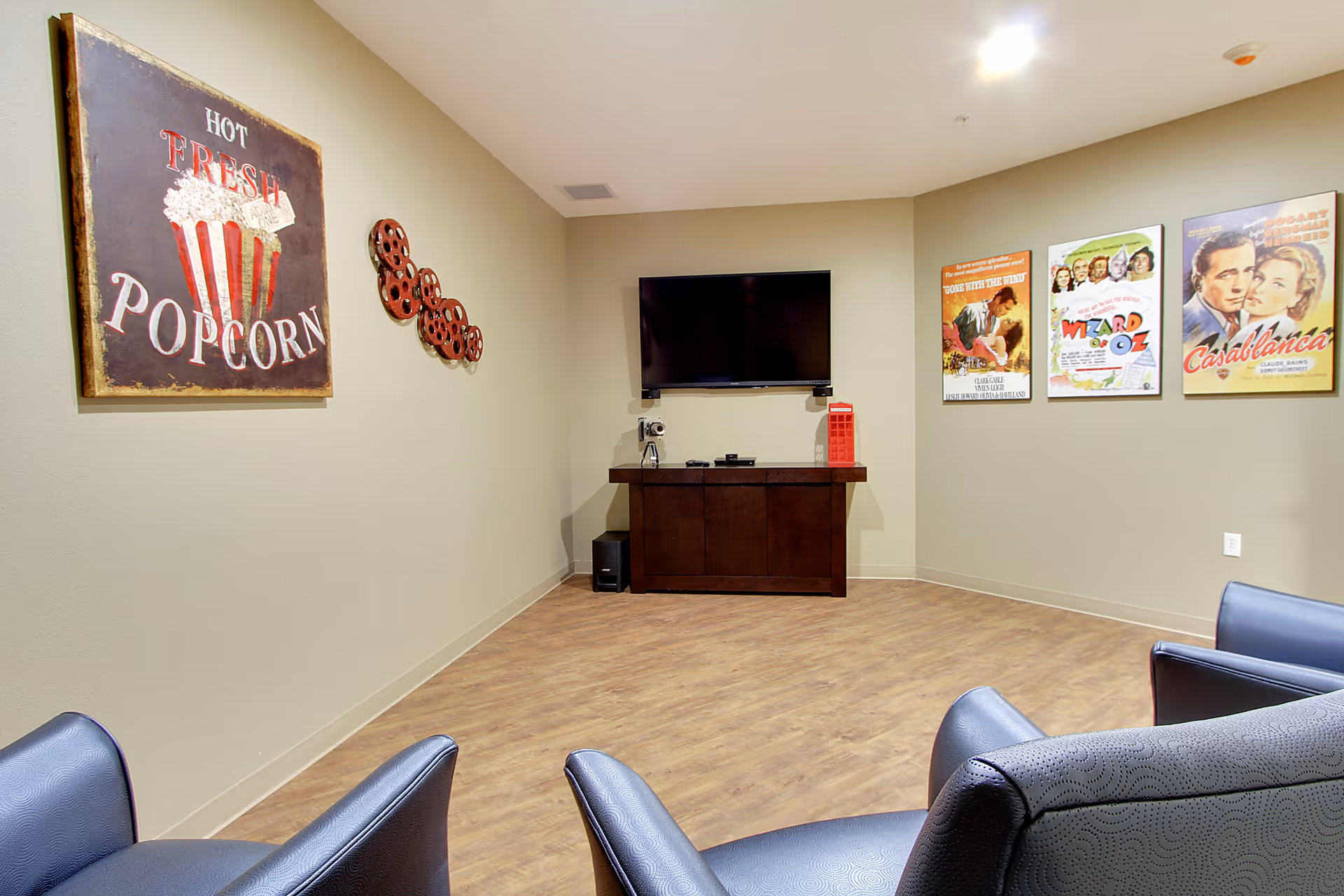 Small media lounge with a wall-mounted TV above a cabinet, movie posters and a popcorn sign on the walls, and several leather chairs.