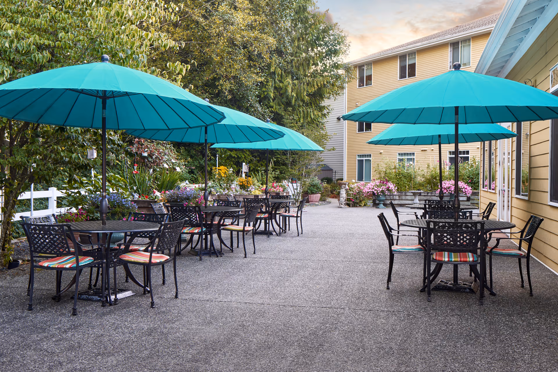 Outdoor patio area at MorningStar Senior Living at Silver Lake with several round metal tables and chairs, each shaded by large teal umbrellas. The patio is surrounded by greenery and colorful flowers, with a yellow building visible on the right side.