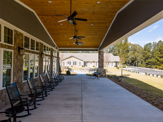 Covered outdoor patio area with a wooden ceiling, ceiling fans, and a row of black rocking chairs along a stone wall with windows. In the background, there is a building with multiple windows and a parking area surrounded by trees and landscaping.