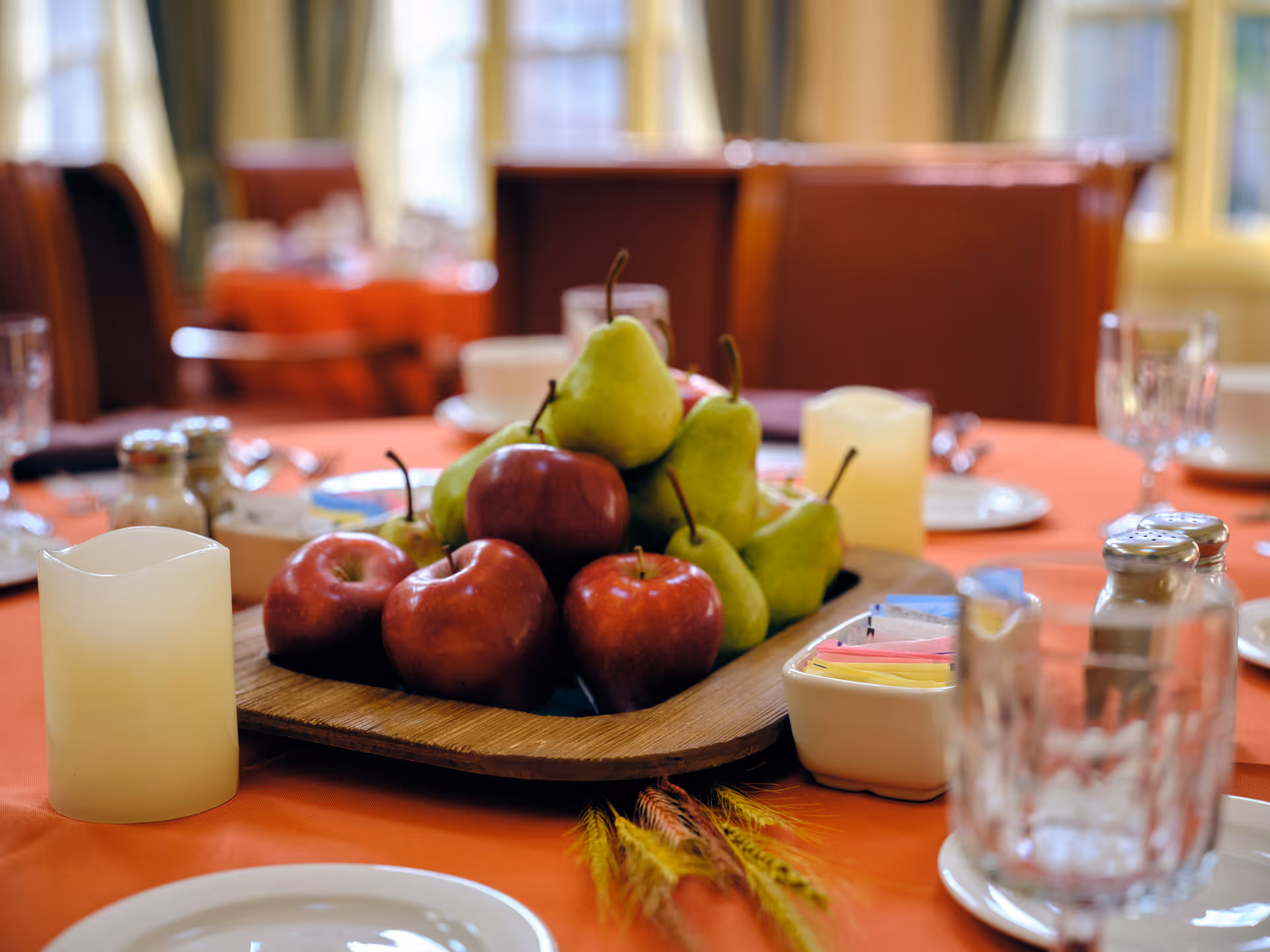 A dining table set with an orange tablecloth, featuring a wooden tray centerpiece filled with red apples and green pears. The table also has salt and pepper shakers, a container with sugar packets, glassware, plates, and a candle. In the background, there are chairs and windows letting in natural light.