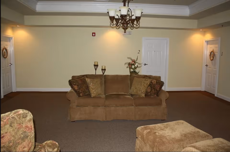 A cozy living room area with a brown sofa adorned with patterned cushions, a matching armchair and ottoman, beige walls with three white doors numbered 1 and 2, a chandelier hanging from the ceiling, and decorative candle holders and a flower arrangement behind the sofa.