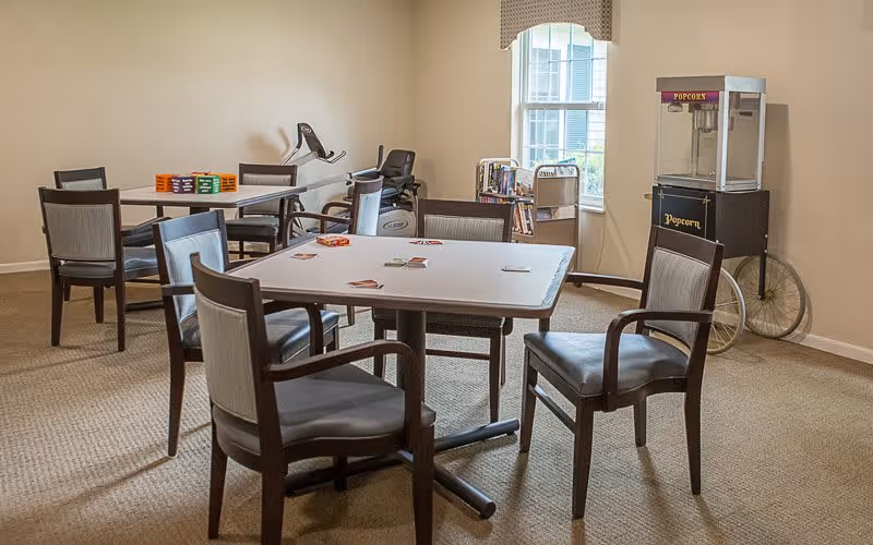 A common area in a senior living facility with two tables and several chairs arranged around them. One table has playing cards on it, and the other has colorful board games. In the background, there is a popcorn machine on wheels and a small bookshelf near a window with beige curtains. The room has beige walls and carpeted flooring.
