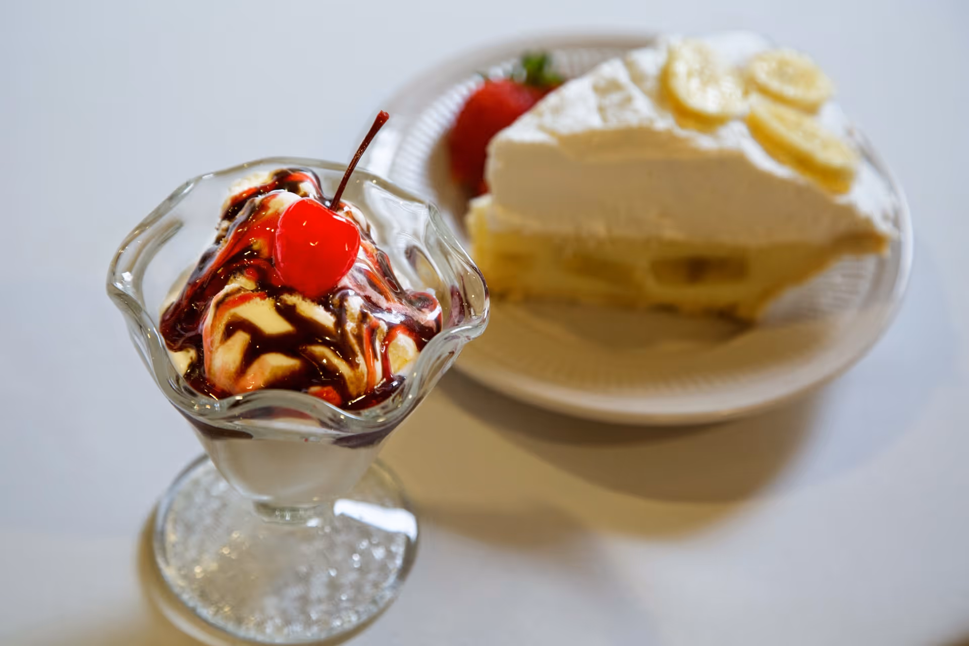A glass ice cream sundae topped with chocolate sauce and a cherry, with a slice of cream pie and a strawberry on a plate in the background.