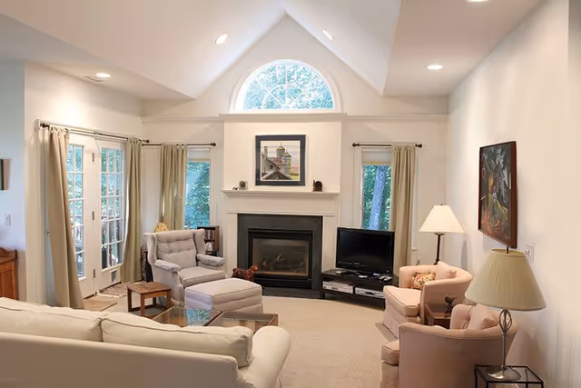 A cozy living room with a vaulted ceiling and a large arched window above a fireplace. The room features a beige sofa, two armchairs, a glass coffee table, a TV on a stand, and lamps providing warm lighting. French doors and windows with curtains allow natural light to enter the space.