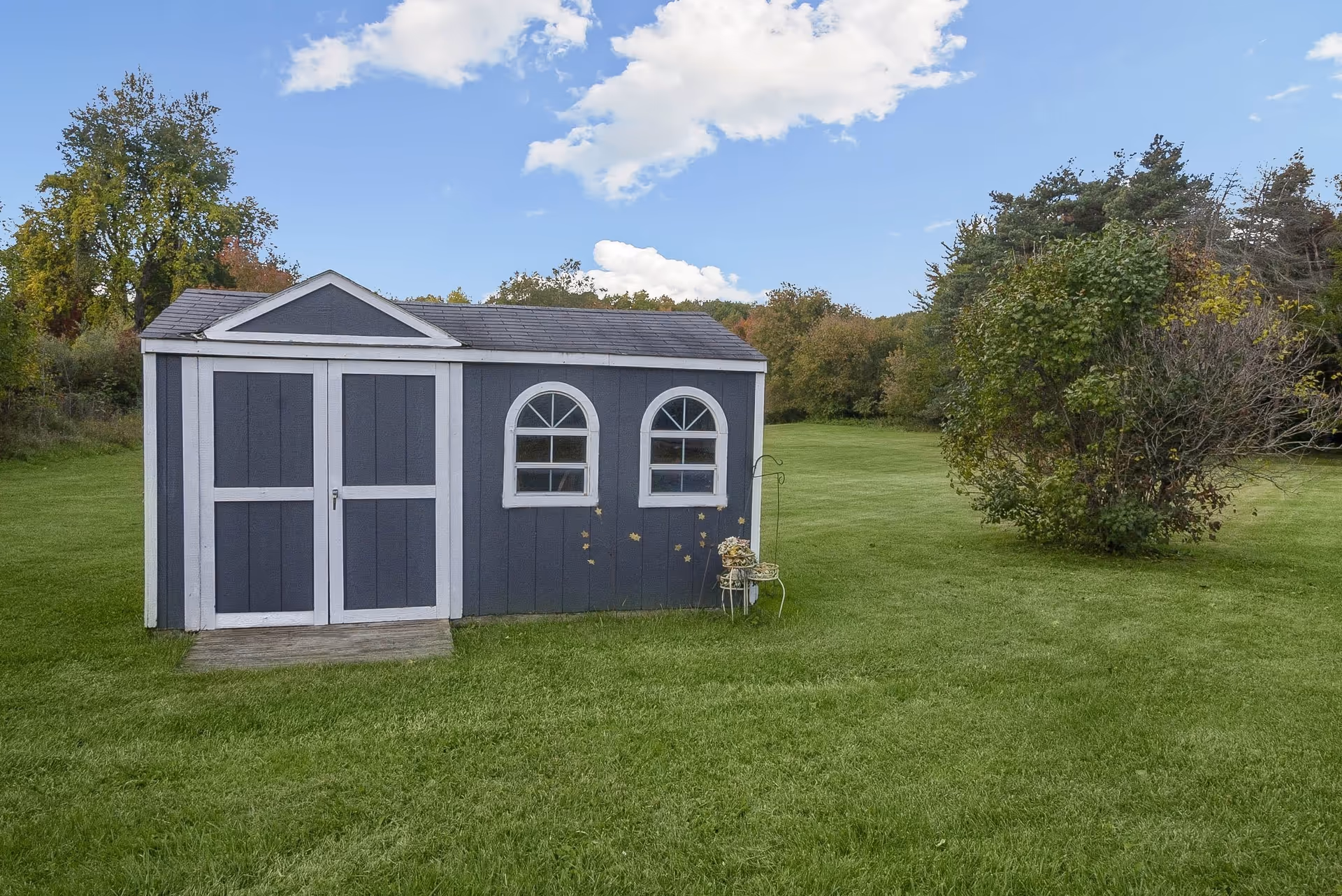 A small gray garden shed with white trim stands on a wide green lawn with trees and a blue sky overhead.