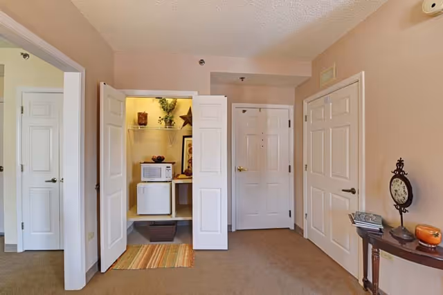 Interior view of a room with beige walls and carpeted floor, featuring three white doors. One door is open revealing a small closet area with a microwave, mini fridge, and decorative items. A small wooden table with a clock and some books is positioned against the right wall.