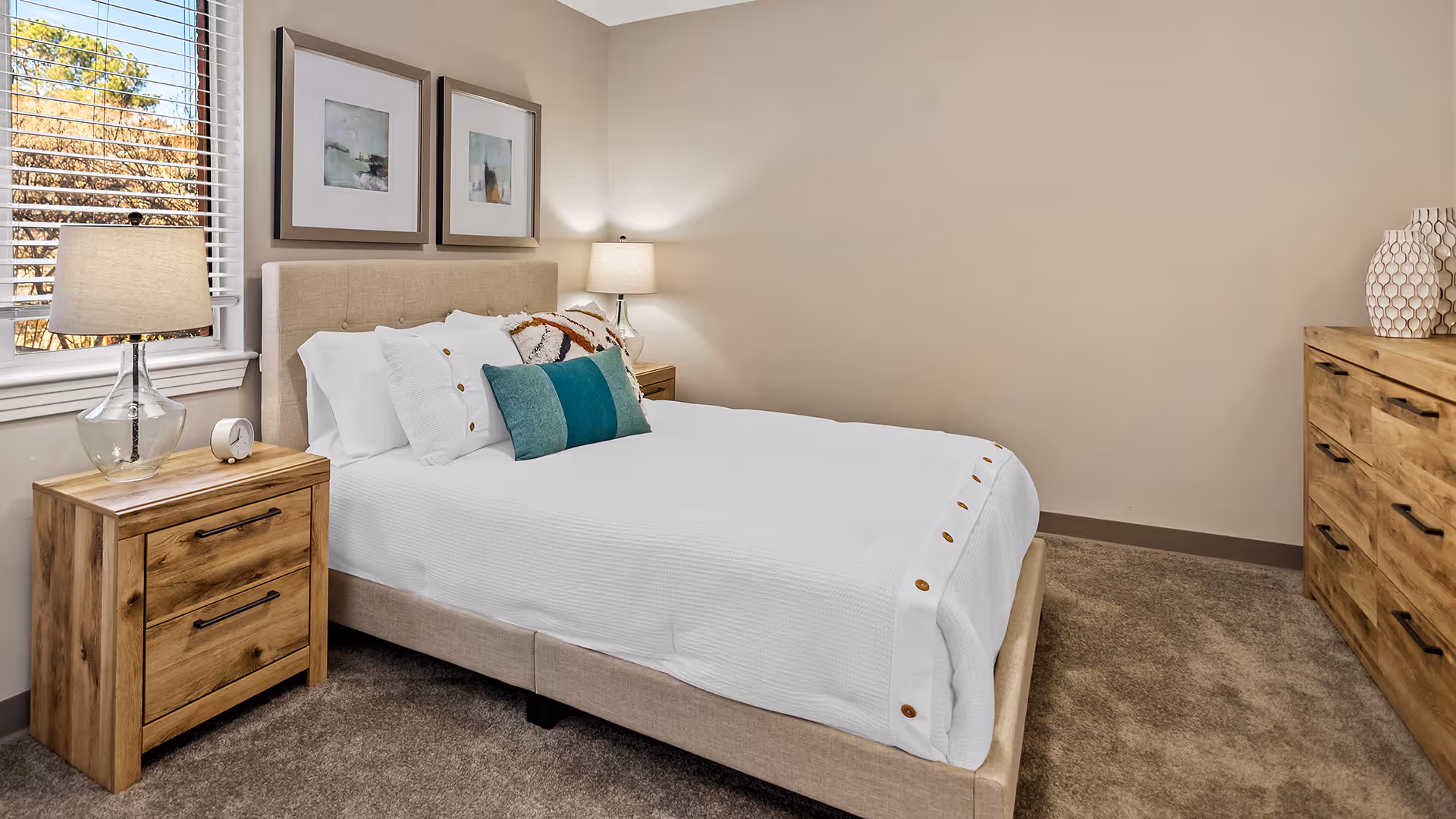A neatly made bed with white bedding and decorative pillows in a bedroom. There are two wooden nightstands with lamps on either side of the bed. A window with blinds is on the left wall, and two framed abstract paintings hang above the bed. A wooden dresser with decorative vases is on the right side of the room. The walls are painted beige and the floor is carpeted.