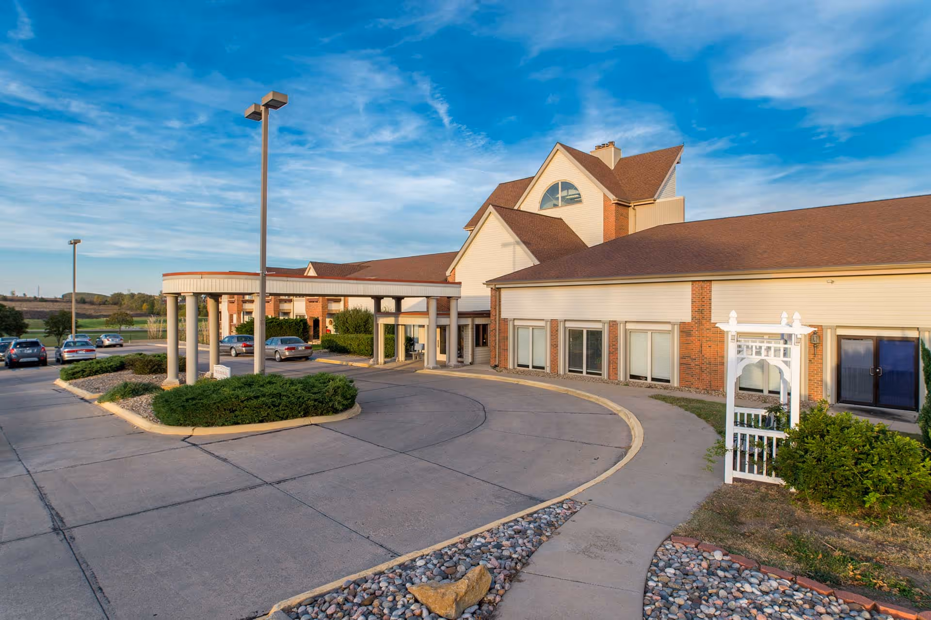 Exterior view of Emporia Presbyterian Manor showing a large building with a covered entrance supported by columns, a curved driveway, landscaped areas with bushes and rocks, several parked cars, and a clear blue sky.