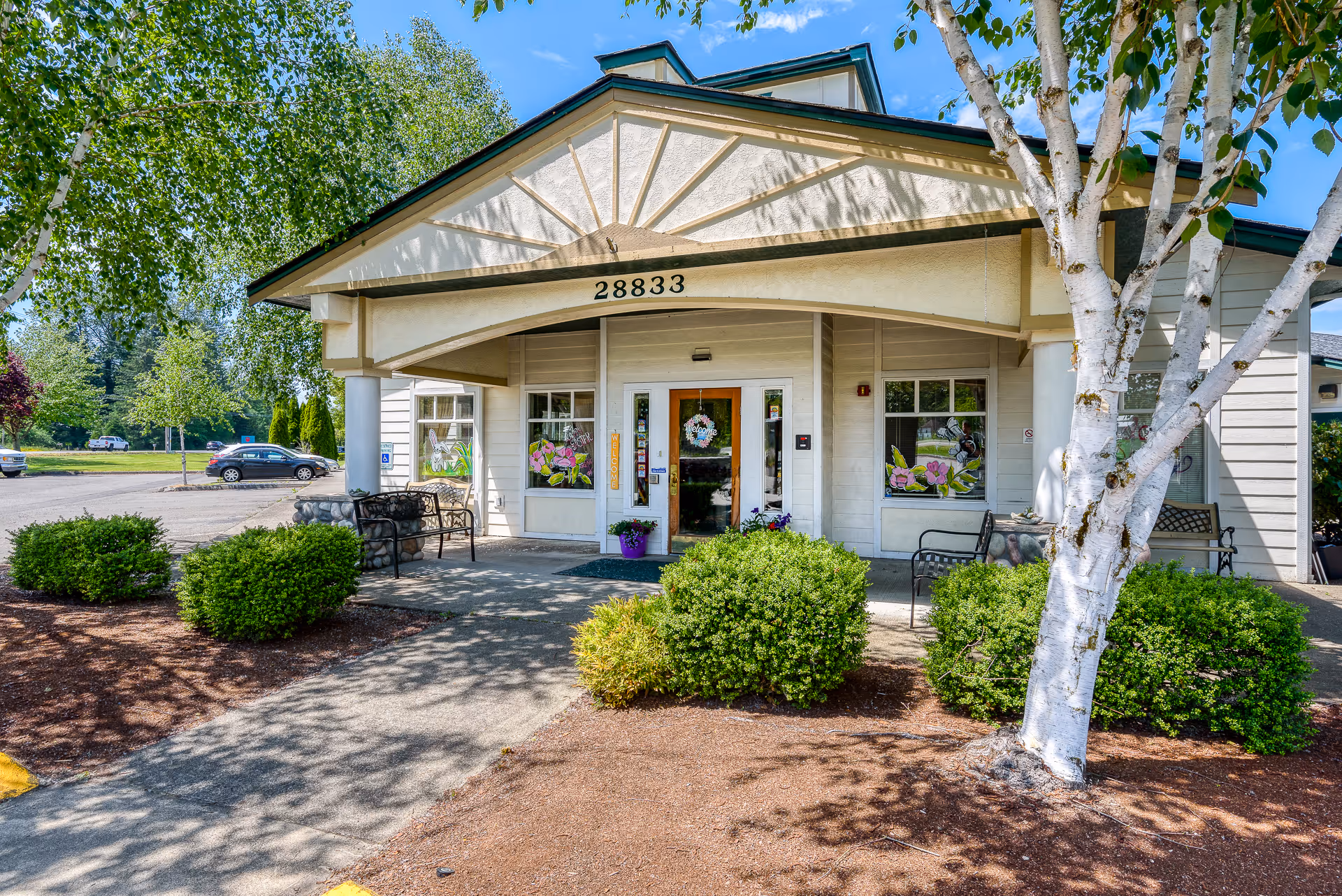 Front entrance of a single-story assisted living building with address '28833', benches, potted plants, and landscaped shrubs.