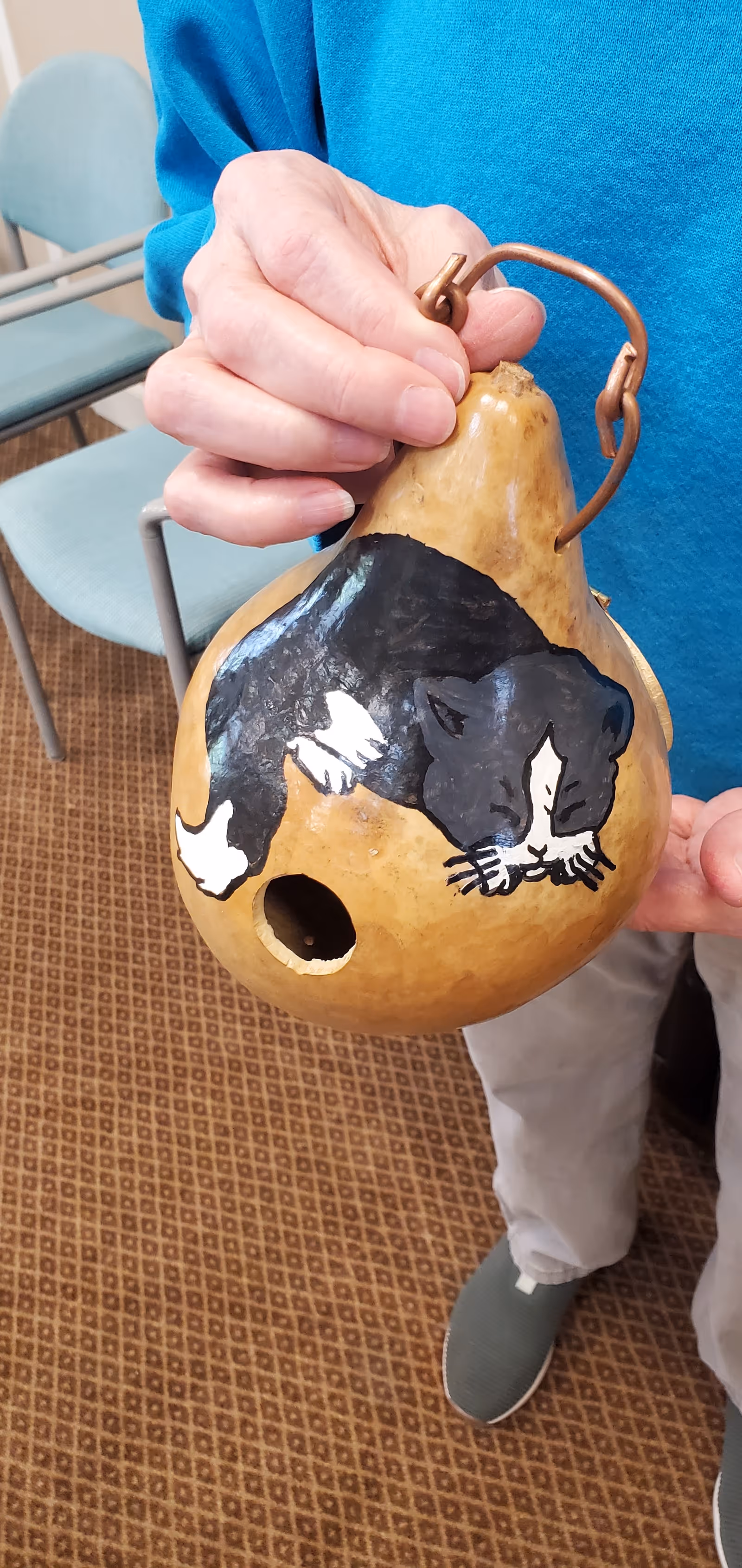 A person holds a painted gourd birdhouse decorated with a sleeping black-and-white cat, with chairs and a carpeted floor visible in the background.