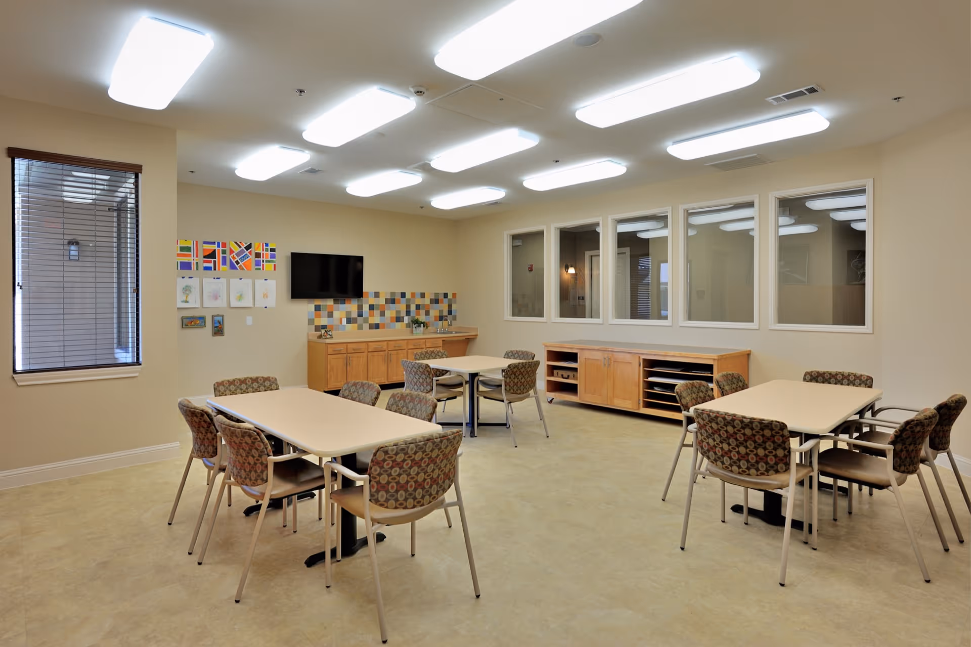A well-lit room with beige walls and floor, featuring three tables each surrounded by four patterned chairs. On one wall, there is a mounted flat-screen TV above a wooden cabinet with colorful square tiles as a backsplash. Several framed artworks and colorful flags are displayed on the wall next to a window with blinds. Another wall has four large windows looking into an adjacent room, and there is a long wooden cabinet beneath these windows.
