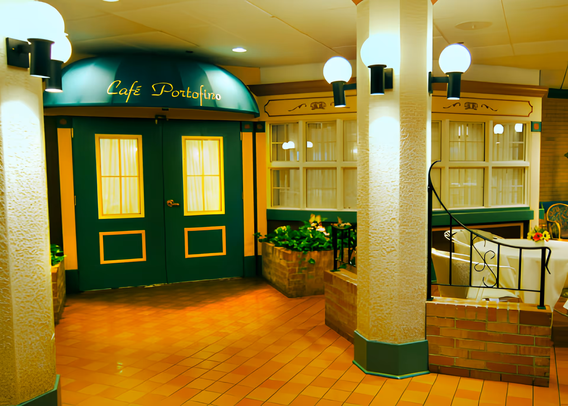 Entrance to Café Portofino inside a facility, featuring green double doors with yellow trim under a green awning with the café name in yellow script. The area has warm lighting, tiled floors, brick planters with green plants, and a seating area with a table and chairs partially visible.