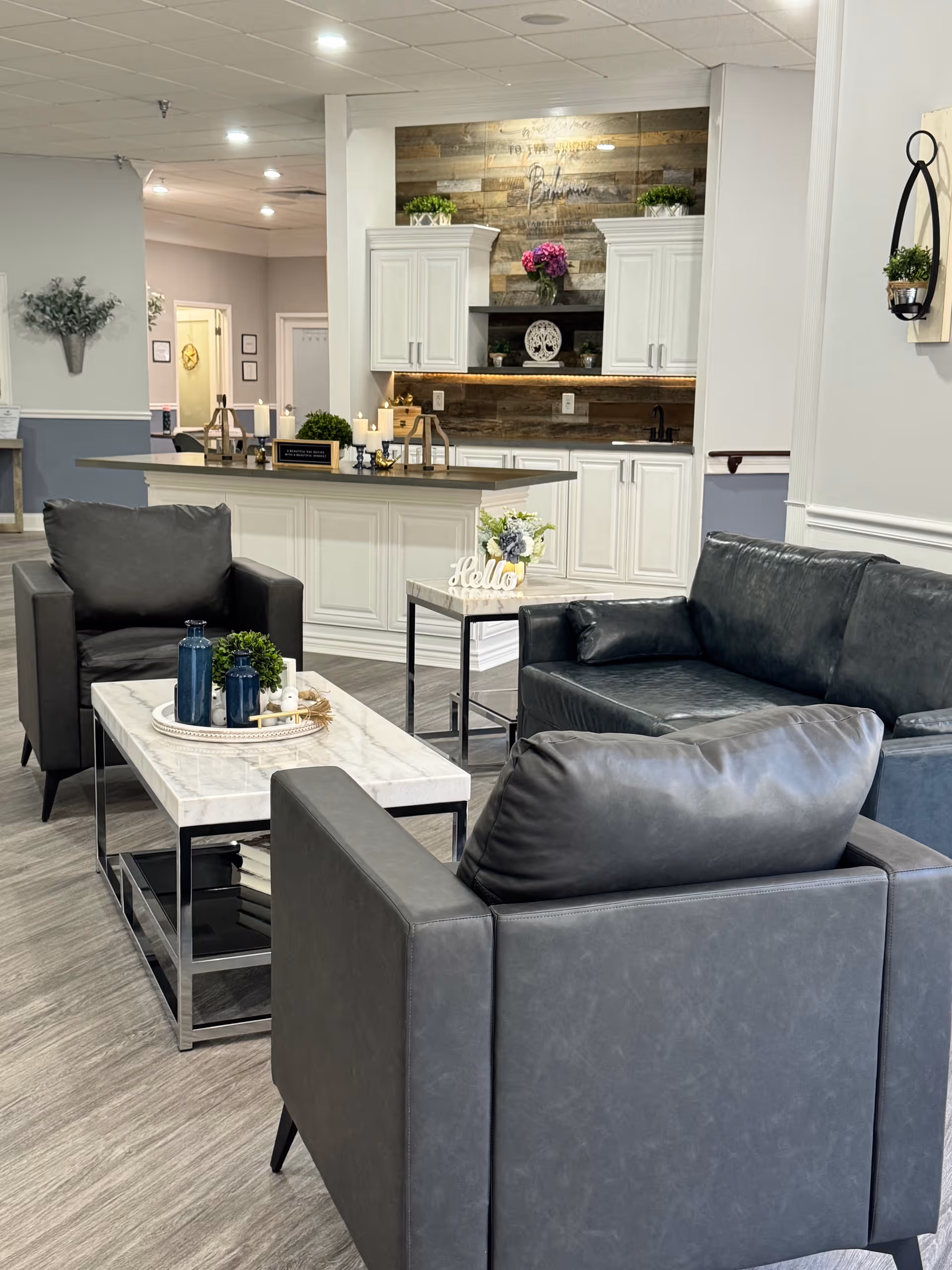 A modern living room area with gray leather sofas and armchairs arranged around a white marble coffee table with decorative items. In the background, there is a white kitchen island and cabinetry with wooden paneling and plants on top. The space is well-lit with ceiling lights and has a welcoming, cozy atmosphere.