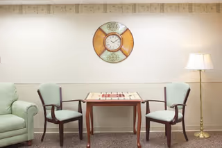 Small sitting area with two upholstered chairs and a game table beneath a decorative wall clock, flanked by a couch and floor lamp.