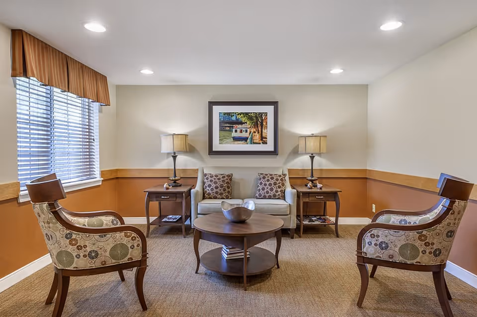 A cozy living room with a beige loveseat adorned with two patterned pillows, flanked by two wooden side tables each with a lamp. Two patterned armchairs face the loveseat across a round wooden coffee table with a decorative bowl. A window with blinds and a valance is on the left wall, and a framed picture hangs on the wall above the loveseat. The walls are painted beige with a brown wainscoting, and the floor is carpeted.