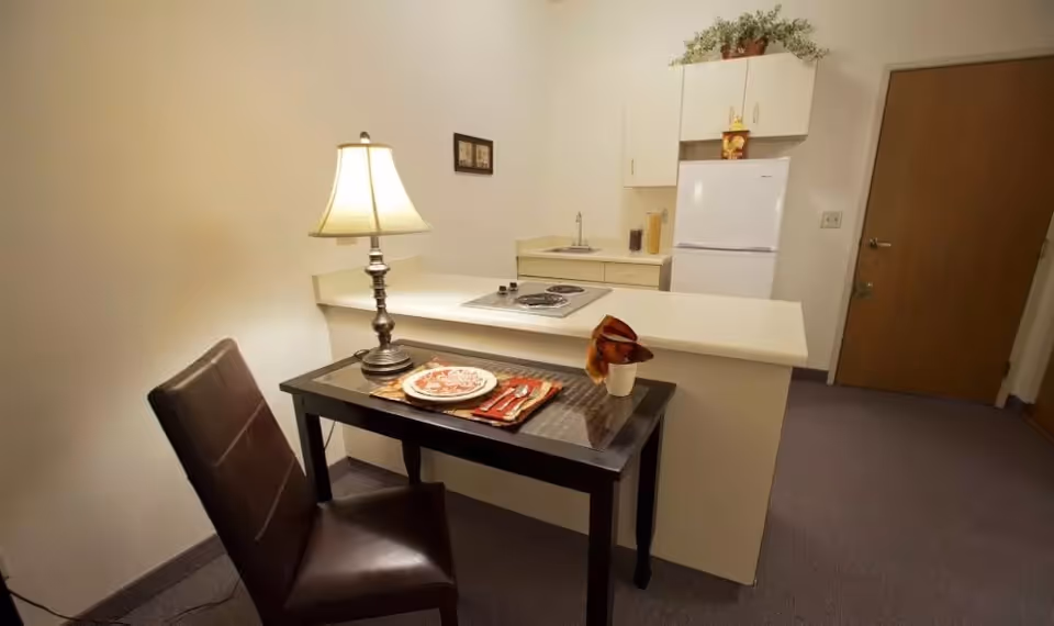 A small kitchen area with a white refrigerator, countertop stove, and sink. In front of the counter is a dark wooden table set with a plate, utensils, napkin, and a lamp. A brown chair is positioned at the table. The room has beige walls and carpeted floor.