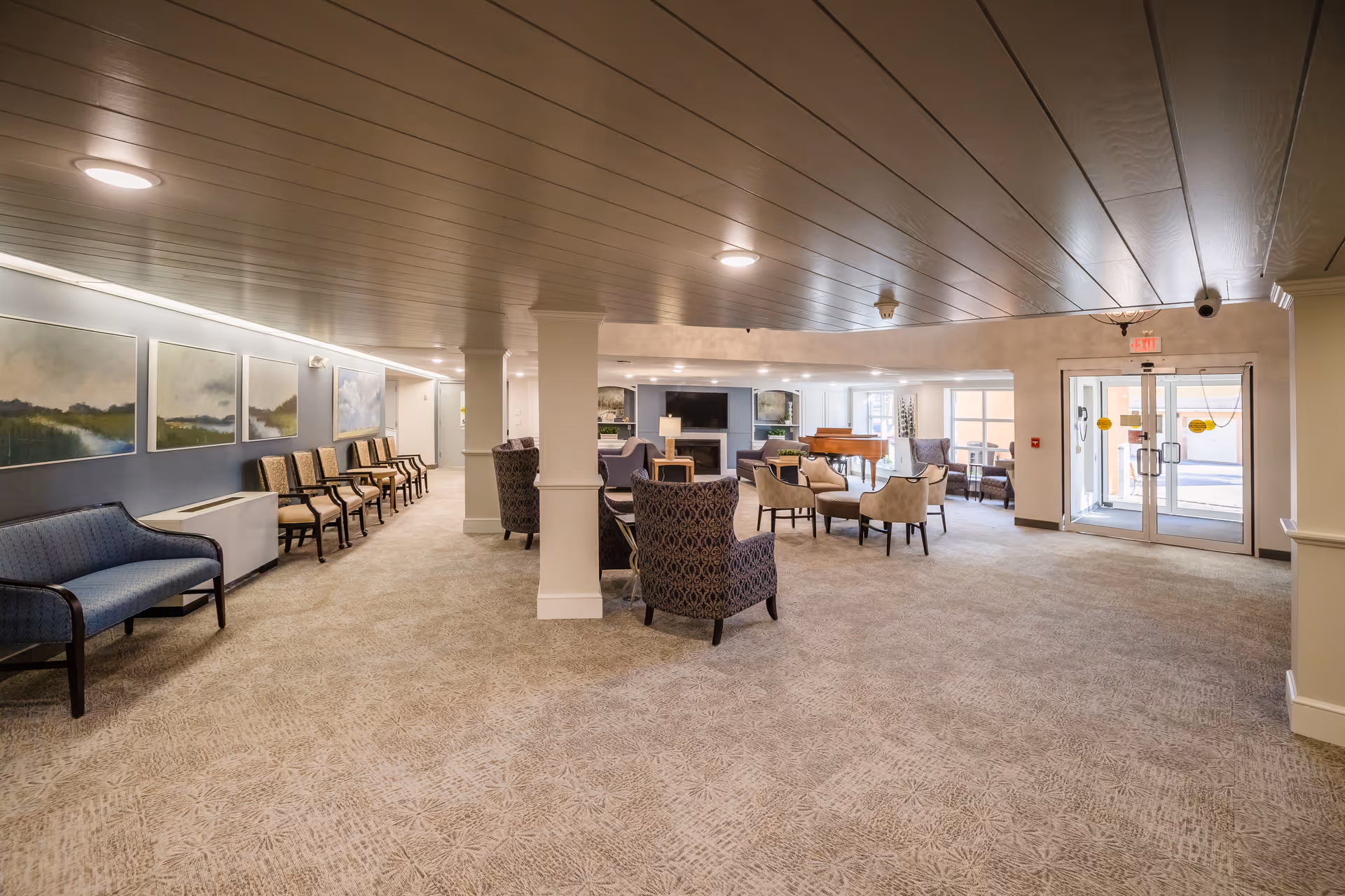 Spacious senior living facility common area with multiple seating arrangements including armchairs and sofas, a piano, and large windows near the entrance door. The room has carpeted floors, a wooden ceiling, and landscape paintings on the wall.