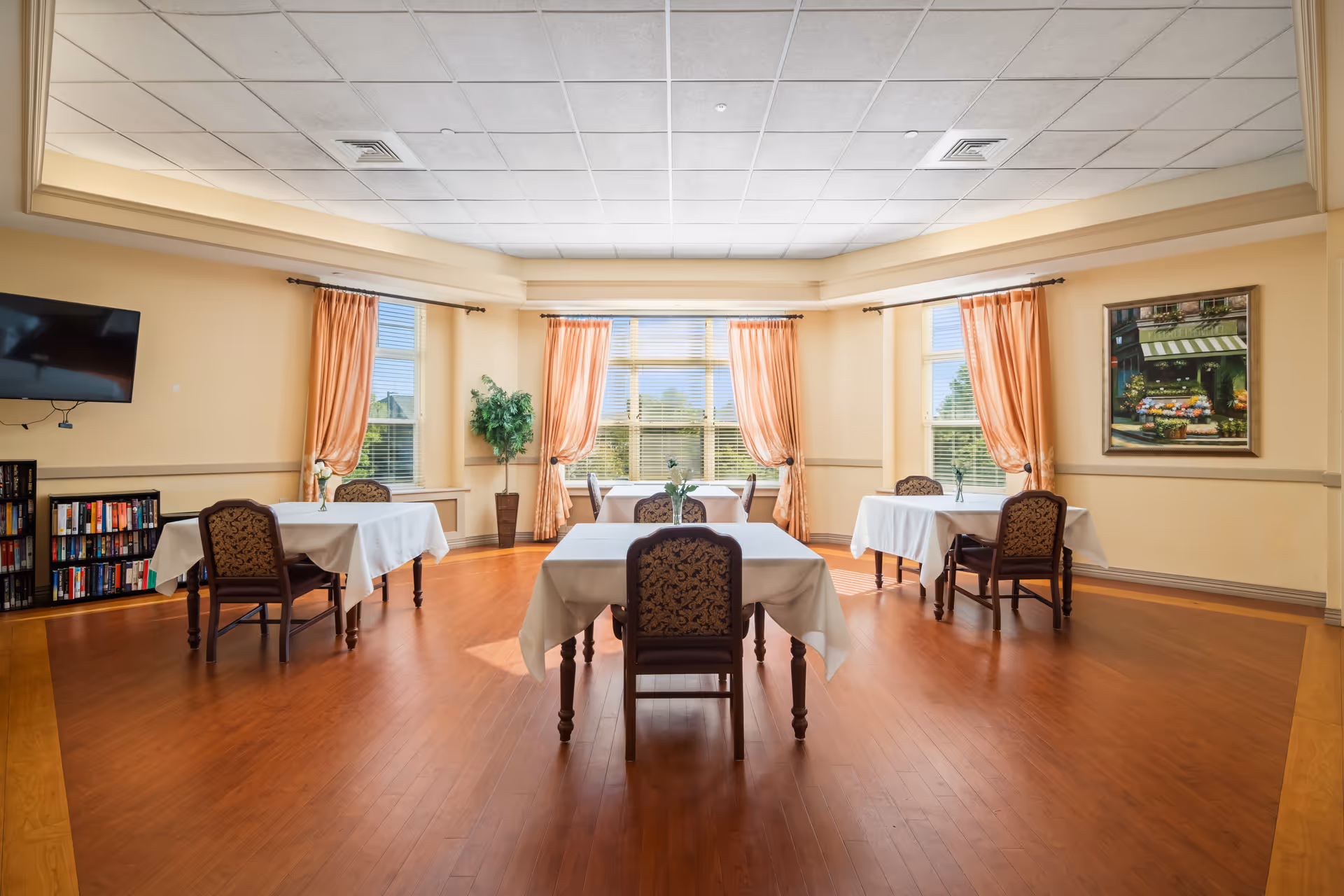 A bright dining room with four tables covered in white tablecloths, each surrounded by chairs with patterned upholstery. The room features large windows with peach-colored curtains, a wooden floor, a flat-screen TV mounted on the wall, a bookshelf filled with books, a potted plant, and a framed painting on the wall.
