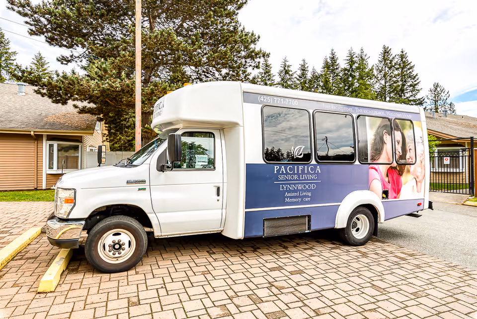 A white and blue shuttle bus parked on a brick driveway near a building. The bus has signage for Pacifica Senior Living Lynnwood, offering assisted living and memory care services. The bus features images of smiling elderly people and a phone number (425) 771-7700.