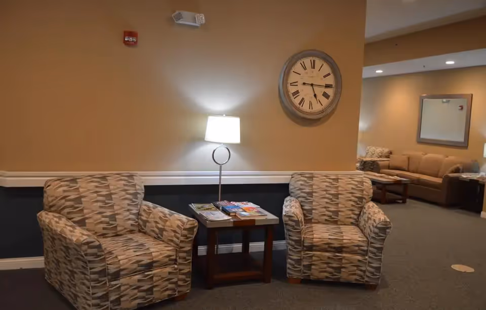 A cozy seating area in a senior living facility with two patterned armchairs separated by a small wooden table holding magazines and a modern lamp. A large round wall clock with Roman numerals hangs above the table. In the background, there is a beige sofa and an armchair with a mirror on the wall.