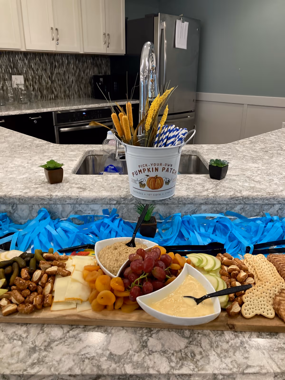 A kitchen countertop with a wooden serving board displaying an assortment of snacks including pretzels, cheese slices, dried apricots, grapes, apple slices, and crackers. Two white bowls contain dips with spoons. Behind the board is a small white bucket labeled 'Pick-Your-Own Pumpkin Patch' holding decorative dried plants and blue-striped straws. The kitchen features a stainless steel refrigerator, a sink with a modern faucet, and white cabinets.