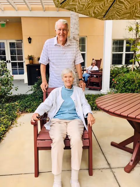An elderly woman sitting in a wooden chair outdoors under a large umbrella, with an elderly man standing behind her. They are in a courtyard area with plants and a building with windows and doors in the background. Another elderly person is sitting in a wooden chair further back near the building.