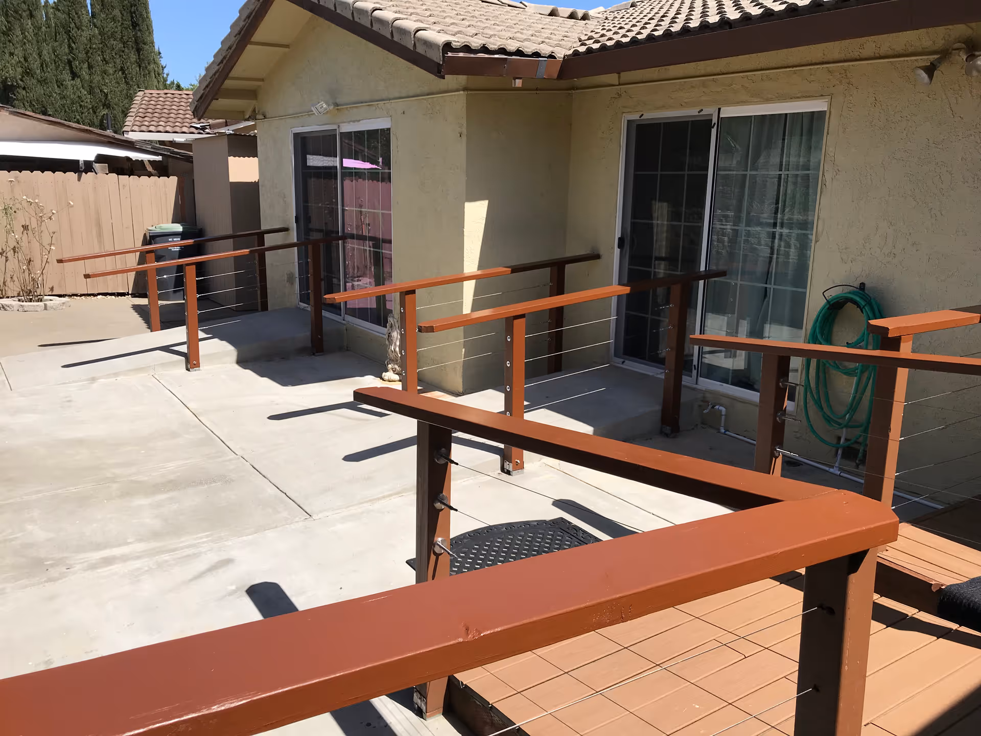 Outdoor patio area of a residential building with beige stucco walls and a tiled roof. The patio has a concrete floor and wooden railings with metal cables. There are two sliding glass doors leading inside, and a green garden hose is coiled on the wall to the right. A wooden fence and some trees are visible in the background.