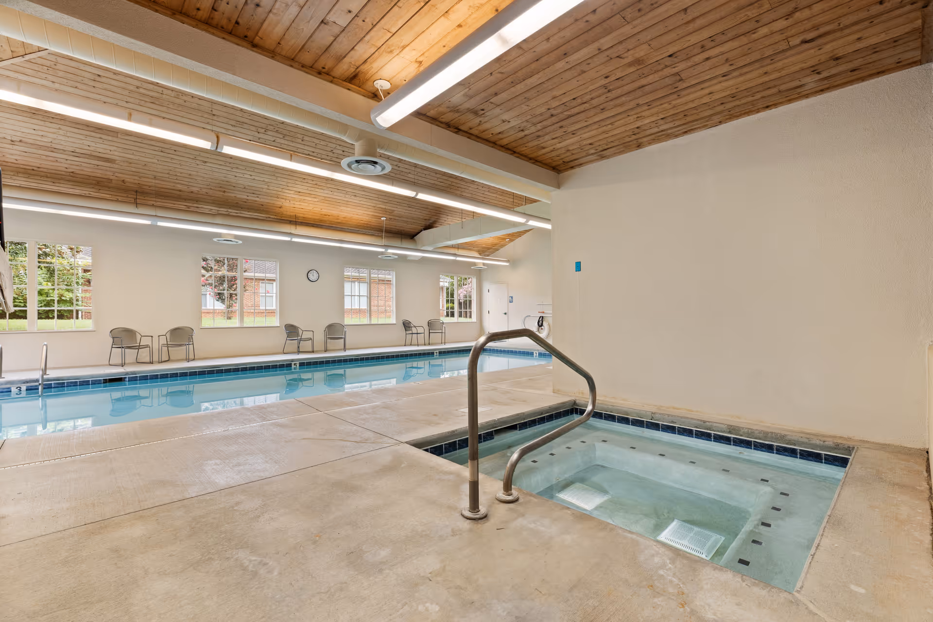 Indoor swimming pool area with a hot tub in the foreground, metal handrails, several chairs along the far wall, large windows letting in natural light, and a wooden ceiling with fluorescent lights.