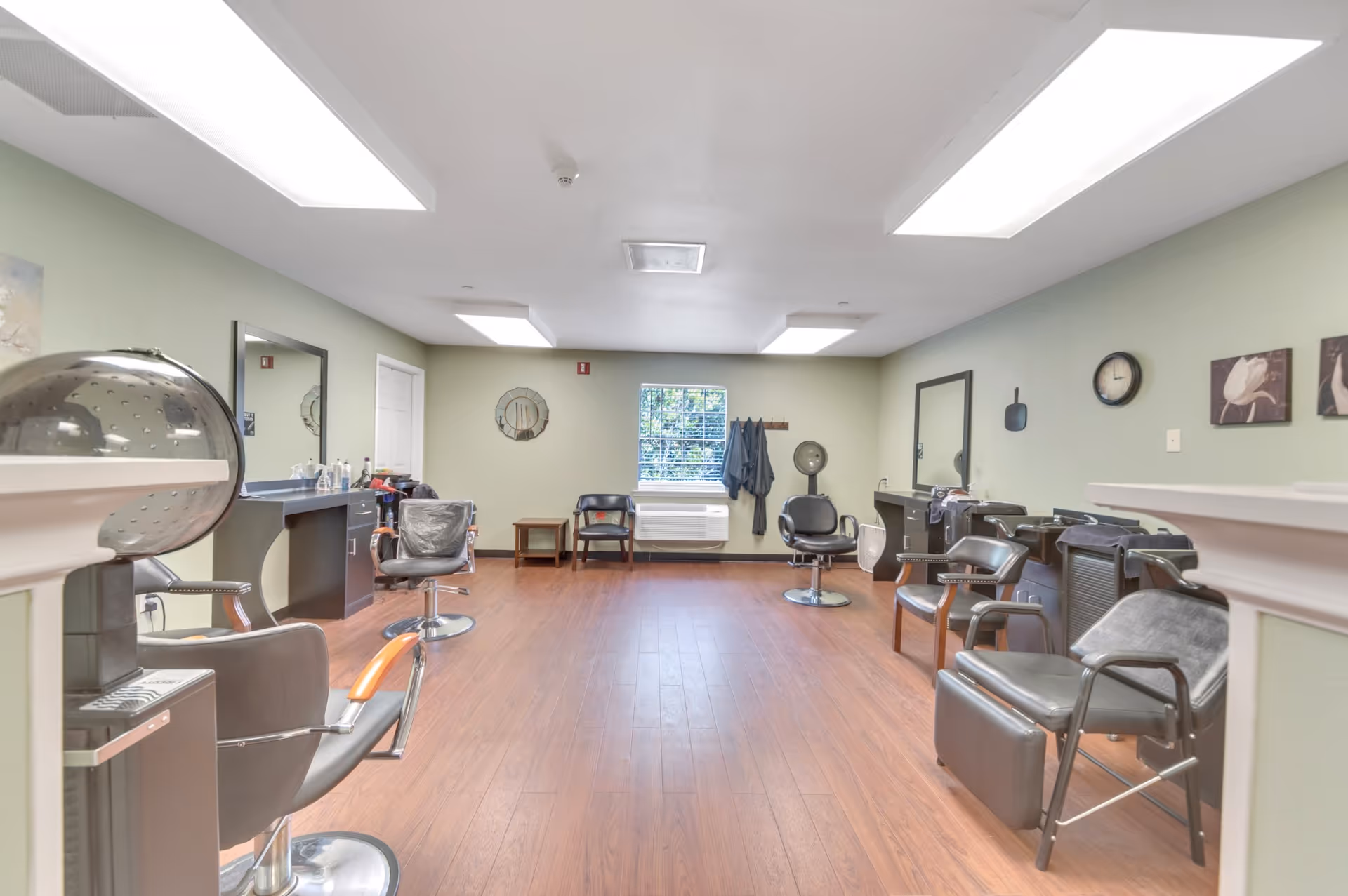 Interior view of a hair salon area in a senior living facility with multiple salon chairs, mirrors, hair dryers, and a window at the far end. The room has light green walls, wooden flooring, and bright overhead lighting.