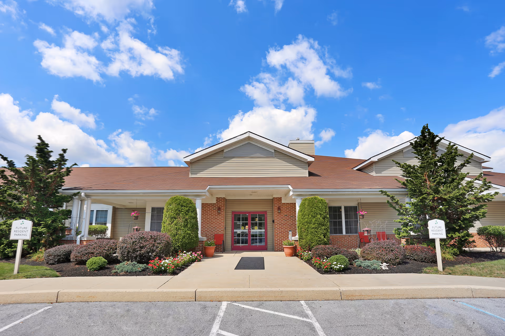 Front entrance of a single-story senior living building with a gabled roof, landscaped beds, and double red doors under a blue sky.
