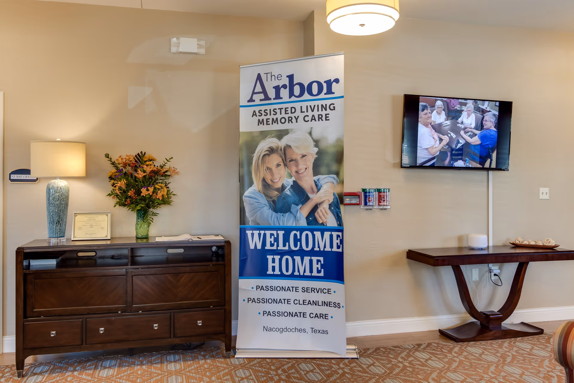 Interior view of a room in The Arbor Assisted Living and Memory Care facility featuring a wooden sideboard with a lamp and a vase of flowers on top, a standing banner with the facility's name and welcome message, and a wall-mounted TV displaying a group of elderly people playing a game.