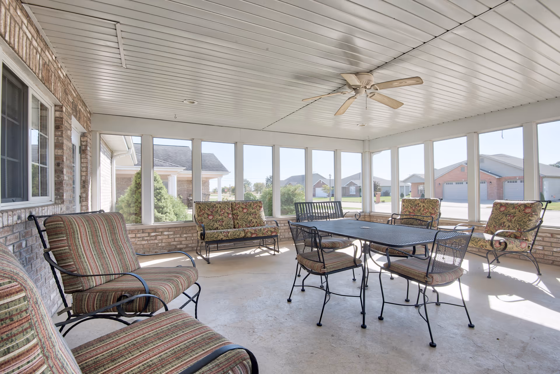 A bright enclosed patio area with large windows on three sides allowing natural light to fill the space. The room features a ceiling fan and several pieces of metal-framed furniture with patterned cushions, including a table with four chairs and multiple armchairs and a loveseat arranged around the perimeter.