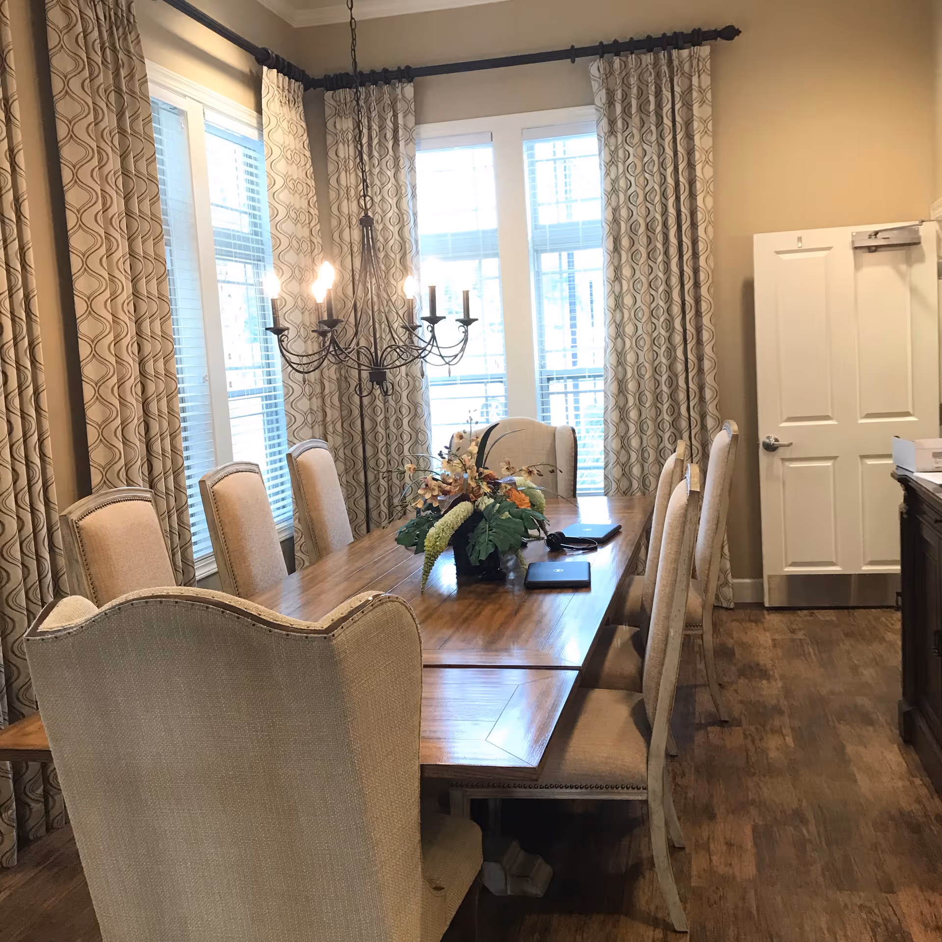 A dining room with a long wooden table surrounded by eight upholstered chairs. A decorative floral centerpiece is on the table. Large windows with patterned curtains allow natural light to fill the room. A chandelier with lit bulbs hangs above the table. There is a white door and wooden flooring.