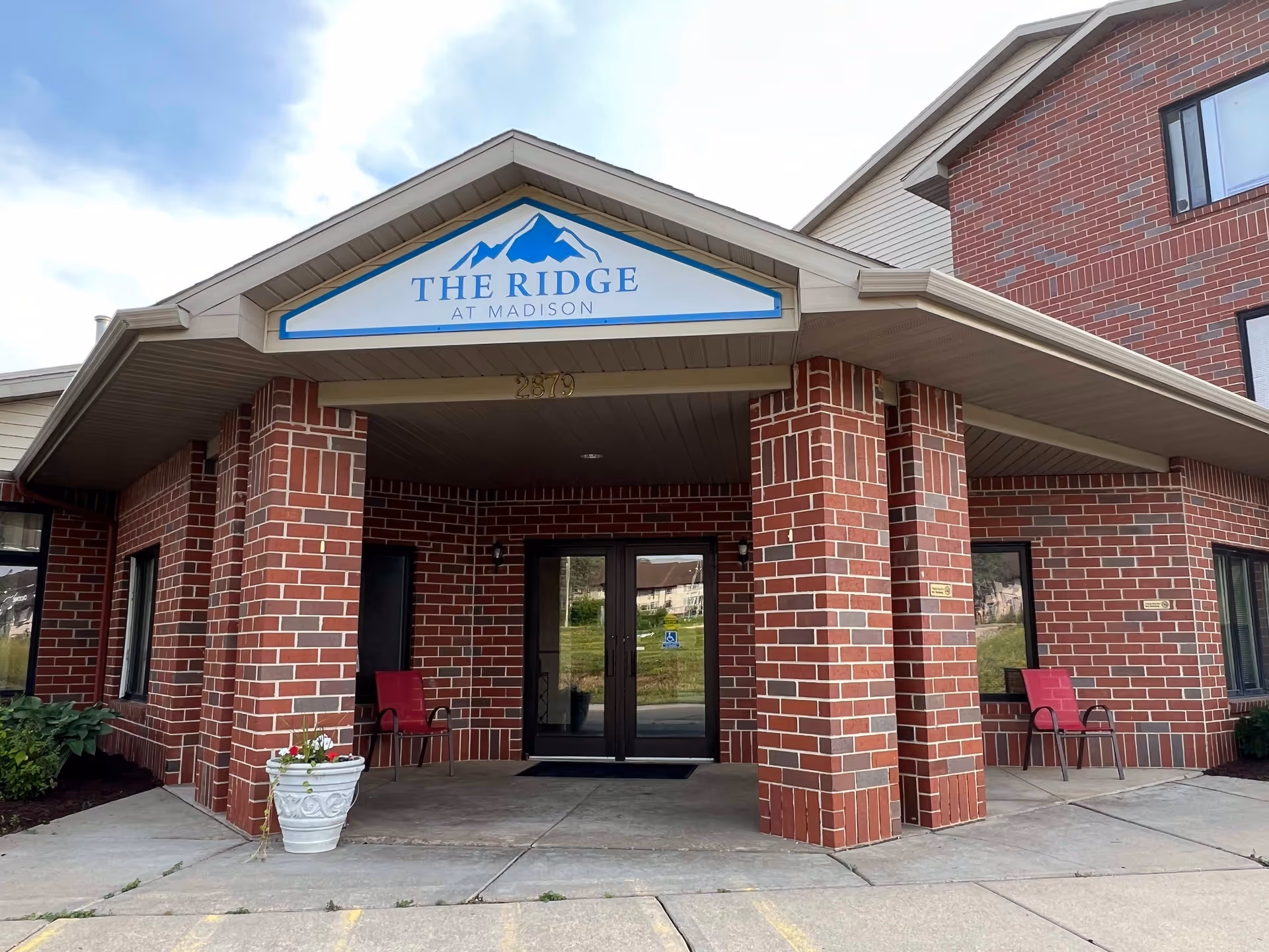 Entrance of The Ridge at Madison facility with a covered porch supported by brick columns, two red chairs on either side of the entrance, a white planter with flowers, and double glass doors.
