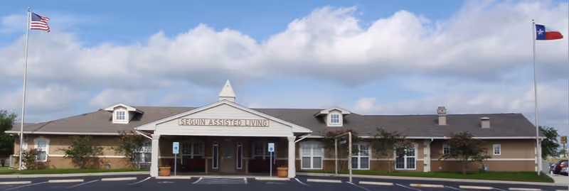 The front exterior of Seguin Assisted Living, a single-story building with a covered entrance flanked by American and Texas flags.