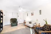 A living room with a white ceiling fan, a green recliner chair, a beige loveseat, a dark patterned armchair, a small wooden side table with a lamp, and a black bookshelf. The walls are white with three framed pictures hanging. The floor is a combination of carpet and tile.