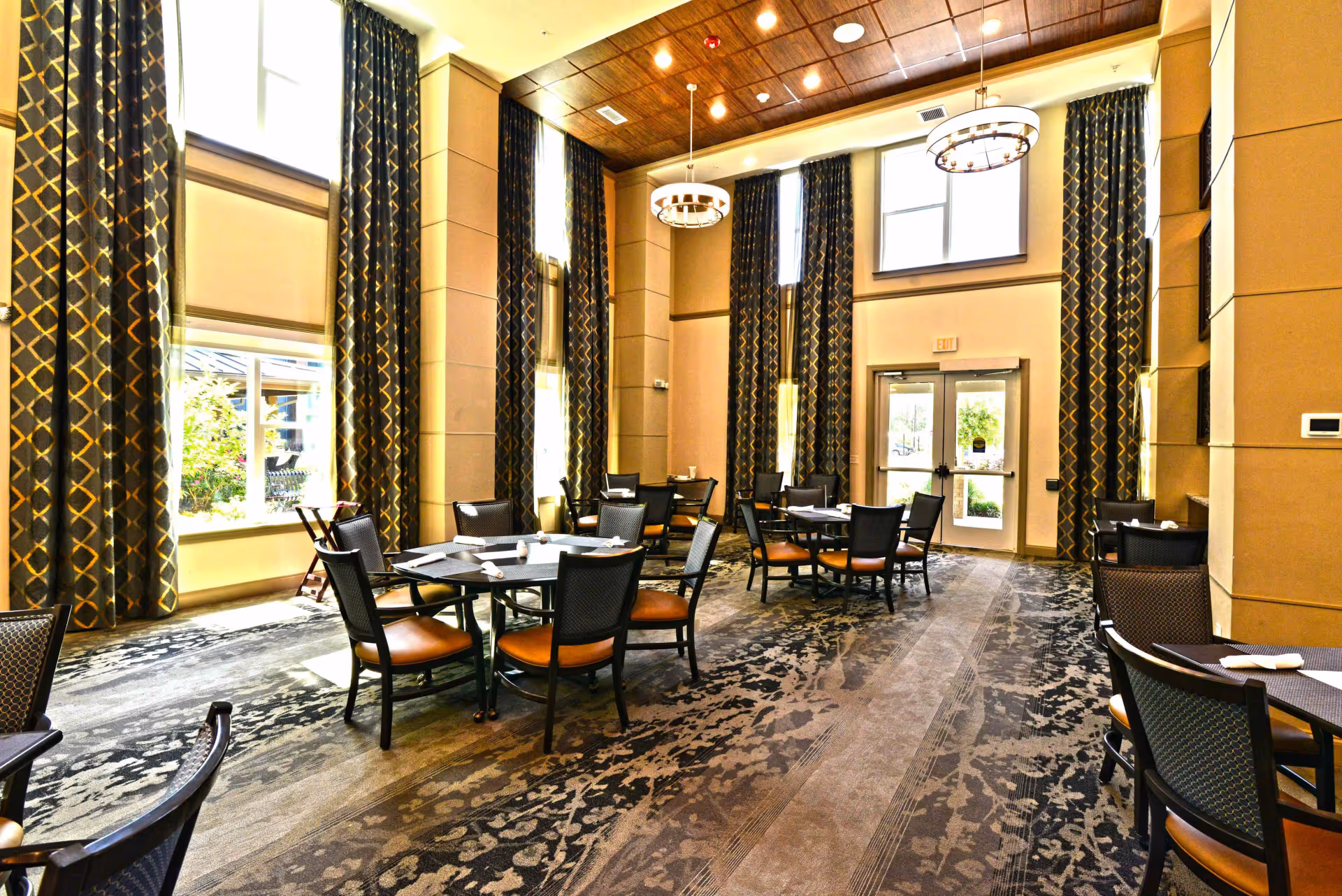 A spacious dining room with multiple round tables and chairs arranged neatly. The room features tall windows with patterned curtains, a high ceiling with recessed lighting and two modern chandeliers. The carpet has a floral and abstract pattern, and there is a glass door leading outside.