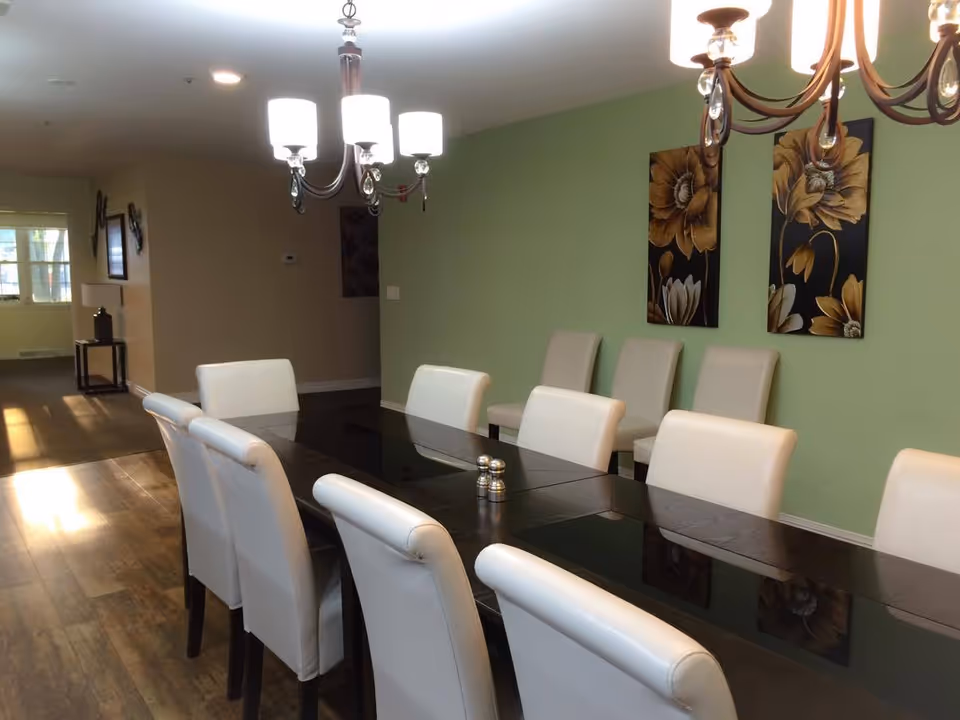 Long dining room with a glossy dark table, white upholstered chairs, chandeliers, and floral wall art on a green accent wall.