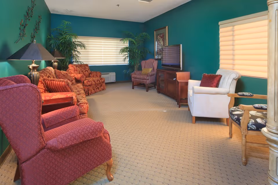 A cozy living room with green walls and beige carpet. The room features multiple upholstered chairs and sofas in red and white tones, a wooden TV stand with a flat-screen television, two large potted plants near the windows with beige blinds, and decorative lamps and wall art.