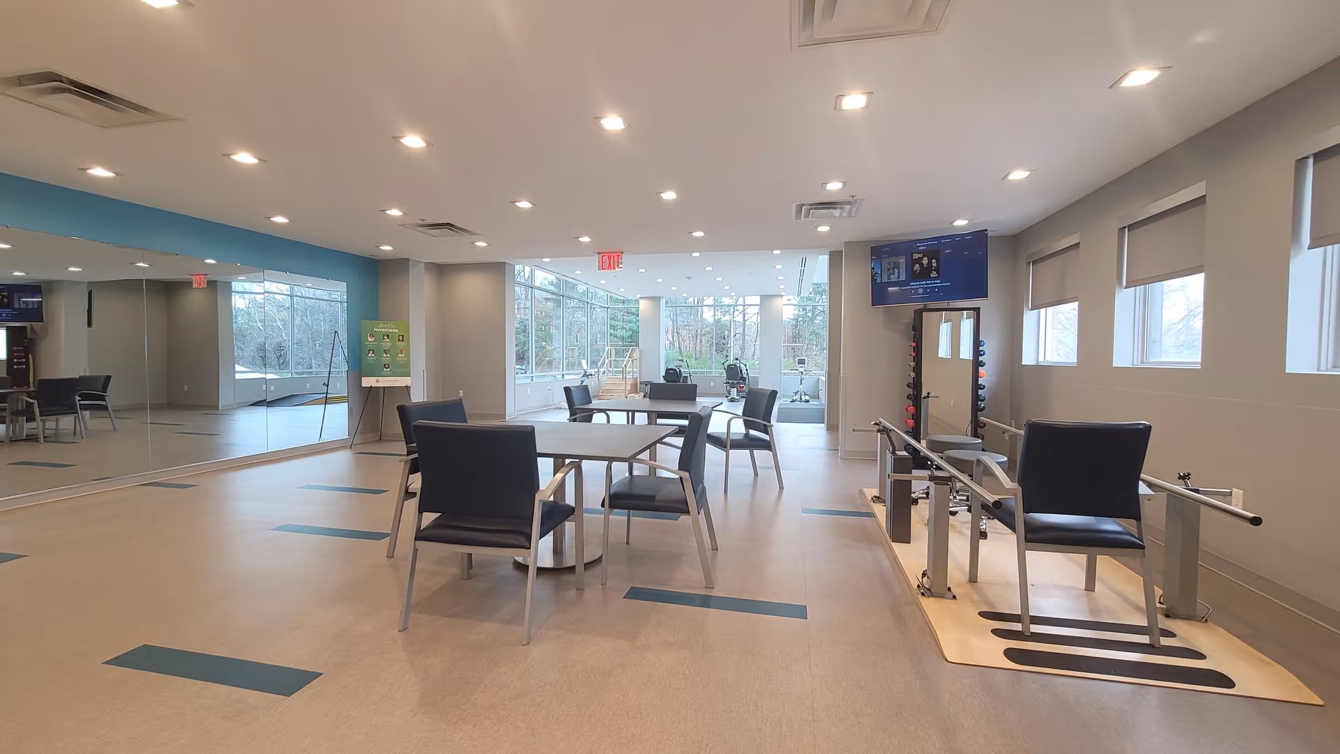 Spacious rehabilitation activity room with tables and chairs, exercise equipment, large windows and a mirrored wall.