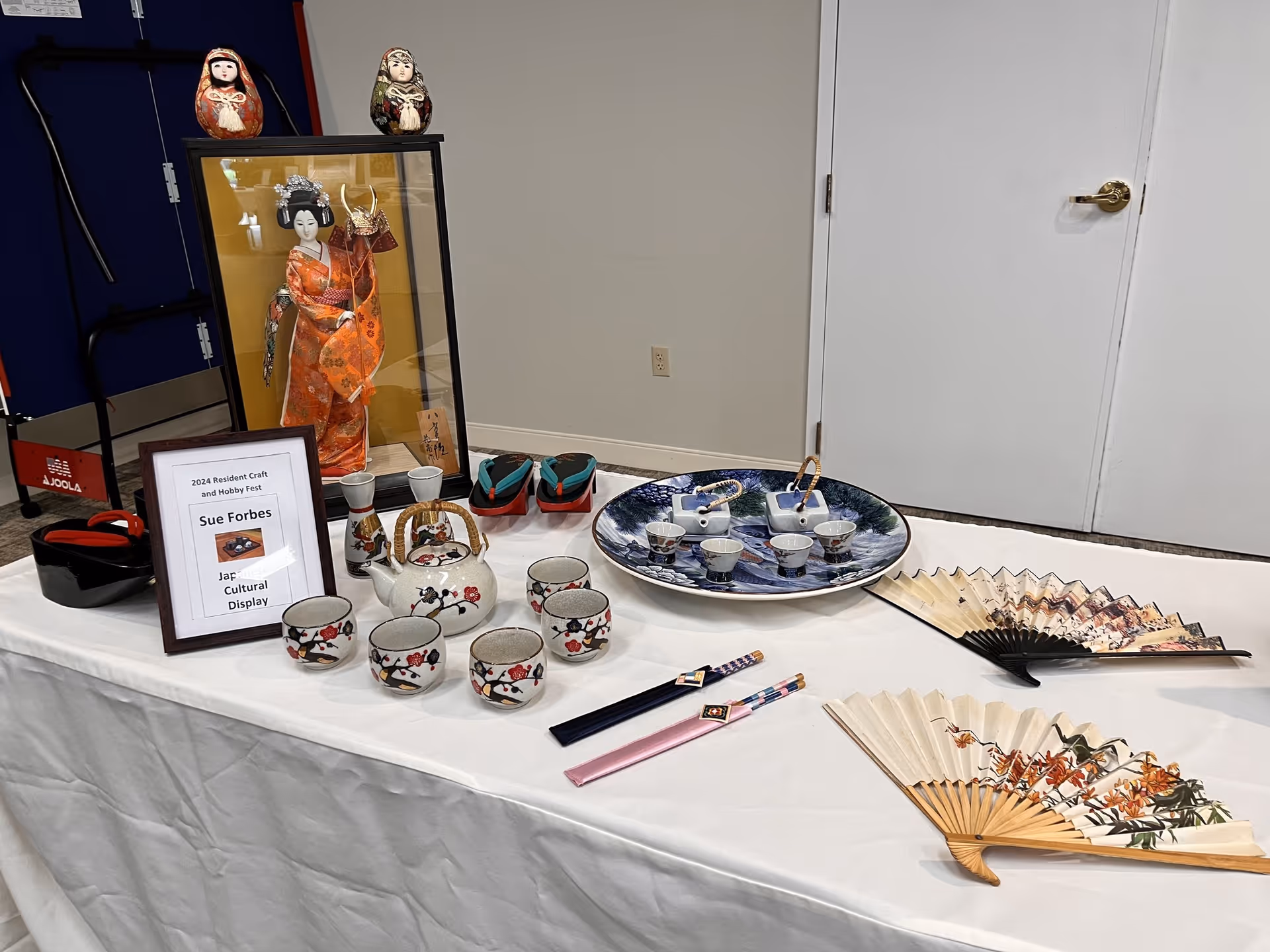A table display featuring Japanese cultural items including a framed picture of a traditional Japanese doll in a glass case, two small dolls, a set of ceramic tea cups and a teapot with floral designs, a large decorative plate with small cups, two folding fans with floral patterns, and a pair of chopsticks. A framed sign on the table reads '2024 Resident Craft and Hobby Fest Sue Forbes Jap Cultural Display.'