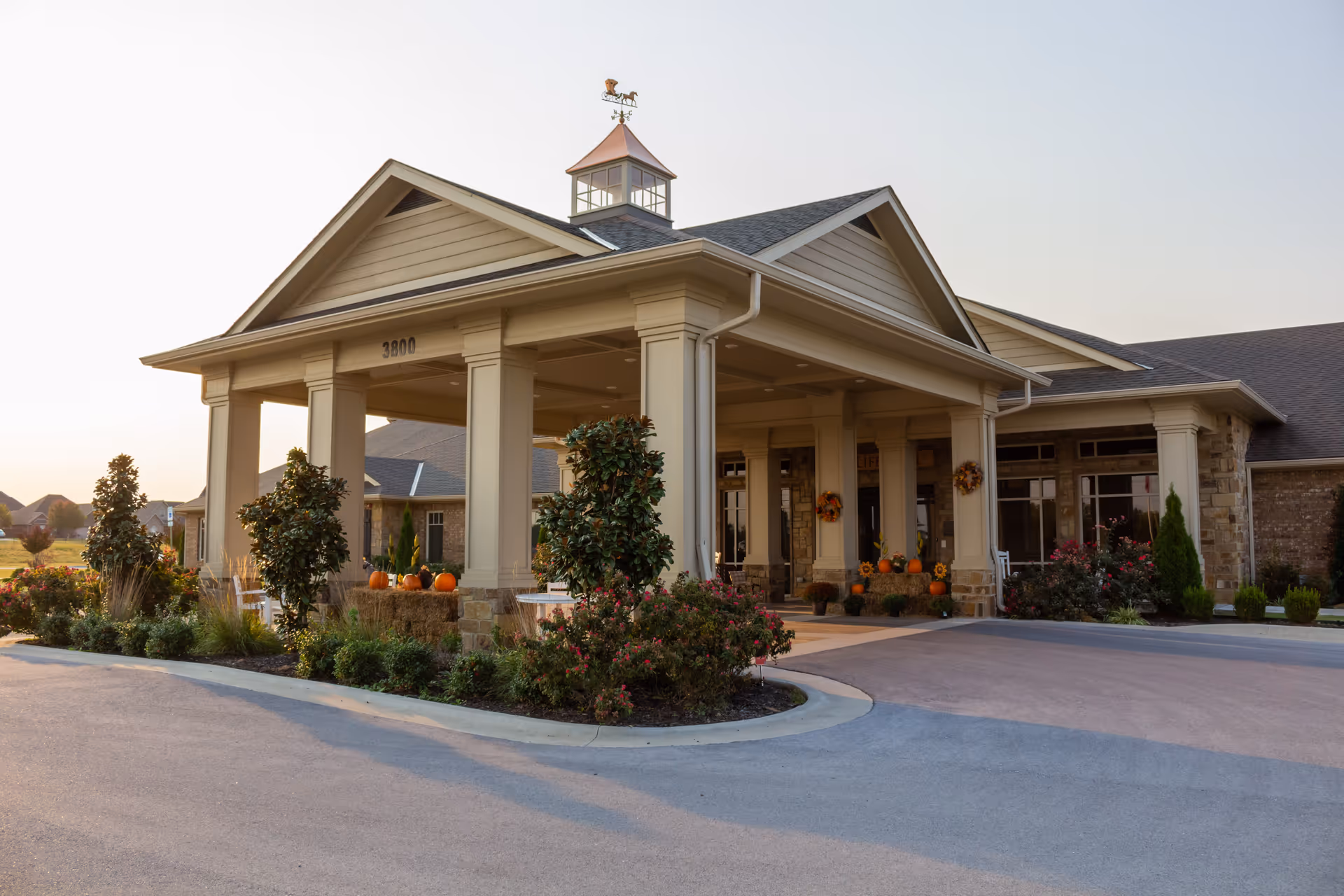 Covered entrance of a senior living facility with columns, landscaped beds and seasonal pumpkin decorations.
