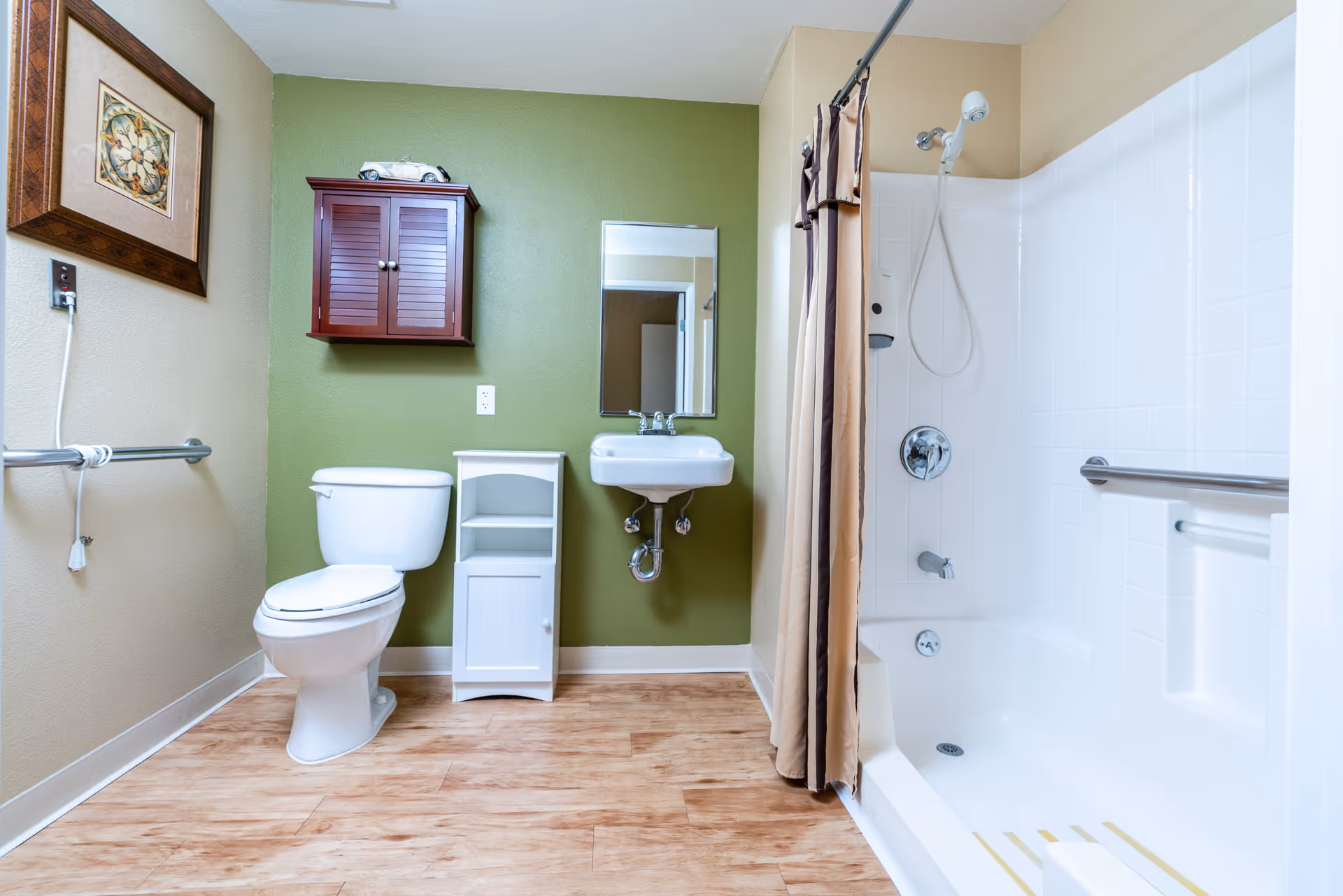 Accessible bathroom featuring a toilet, wall-mounted sink and mirror, storage cabinets, and a bathtub with shower and grab bars.