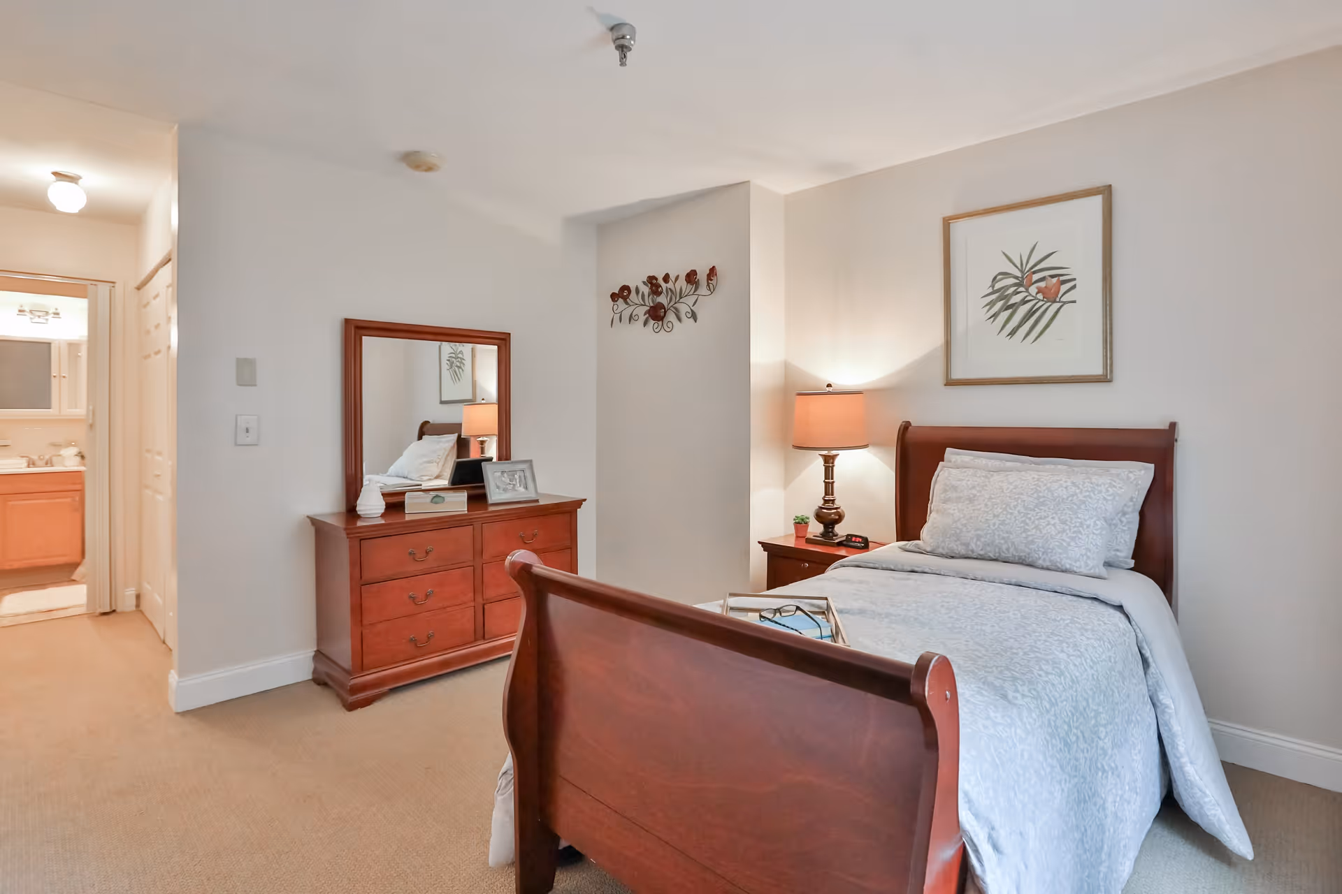 A cozy senior living bedroom featuring a wooden sleigh bed with white bedding and pillows. Next to the bed is a wooden nightstand with a lamp and a small plant. Across from the bed is a wooden dresser with a mirror, decorative items, and a framed photo. The room has beige walls and carpet, with a framed botanical print hanging above the bed. A doorway leads to a bathroom with a vanity and mirror visible.