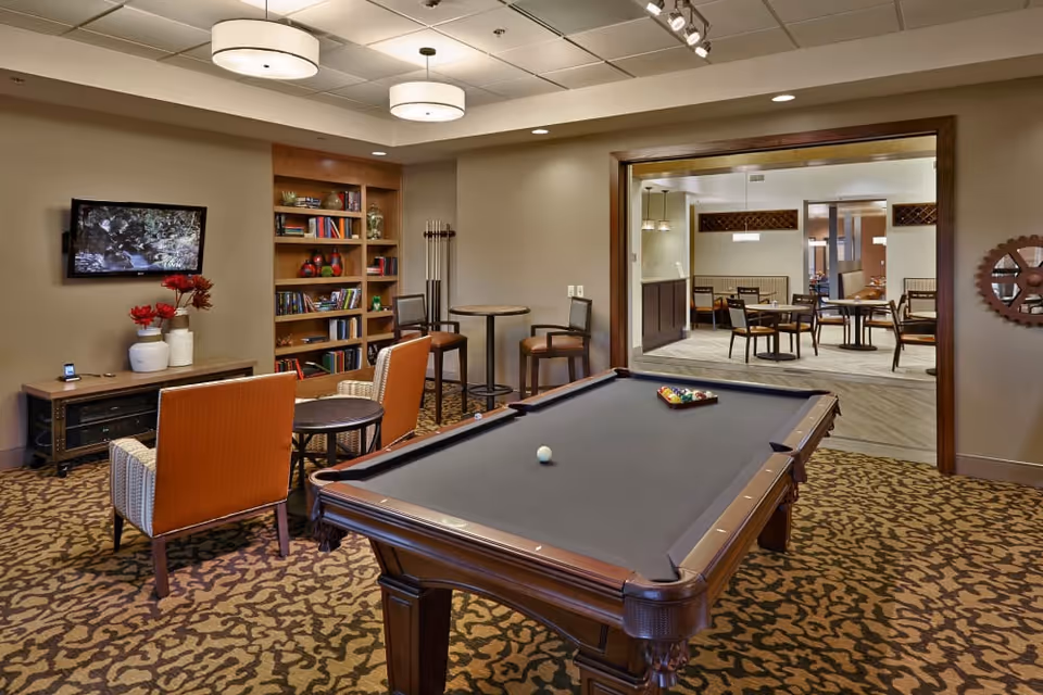A recreational room featuring a pool table with balls arranged for a game, two orange chairs facing a small round table, a wall-mounted TV, a bookshelf with books and decorative items, and a small high table with two chairs. The room opens into a dining area with multiple tables and chairs.