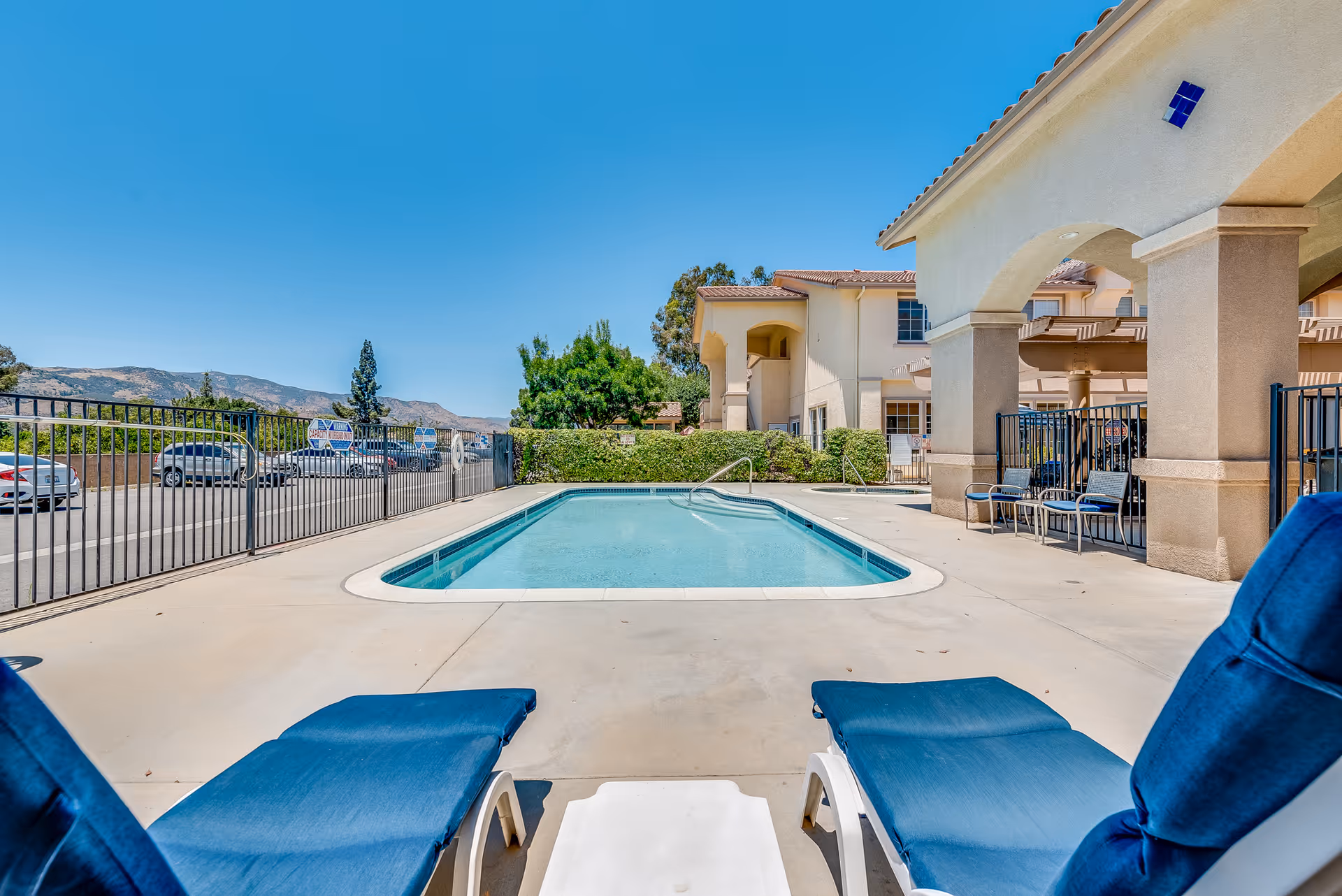 Outdoor swimming pool with two blue lounge chairs in the foreground, a fenced pool deck and the assisted living building with hills in the background.