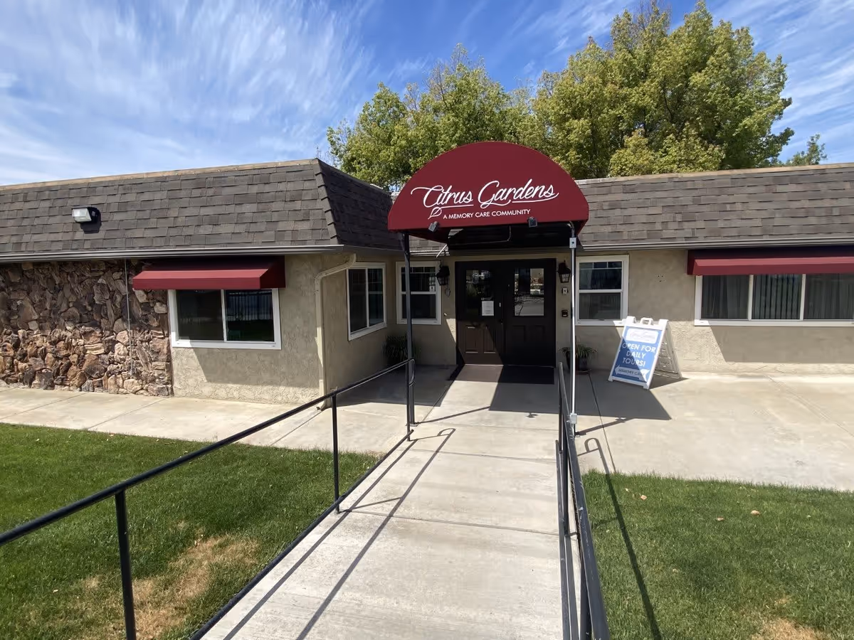 Entrance to Citrus Gardens Memory Care Community with a maroon awning above the door, a concrete ramp with black railings leading to the entrance, beige building exterior with stone accents on the left side, and green grass on both sides of the ramp under a partly cloudy blue sky.