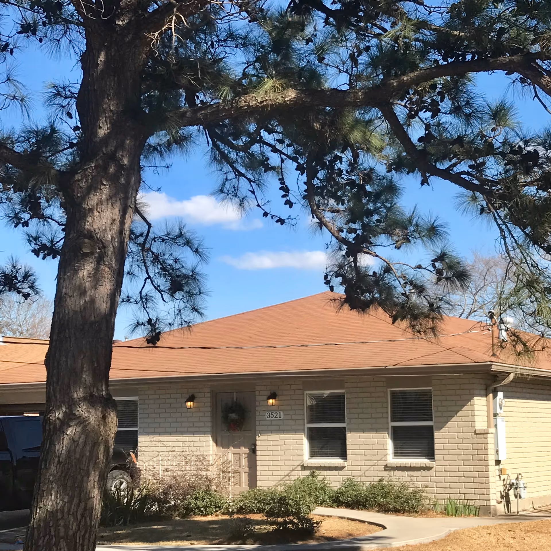 Exterior view of a single-story brick building with a reddish-brown roof, two windows with blinds, a door with a wreath, and two wall-mounted lights on either side of the door. A large tree with pine cones is in the foreground, and a curved concrete walkway leads to the entrance. The sky is clear with a few clouds.