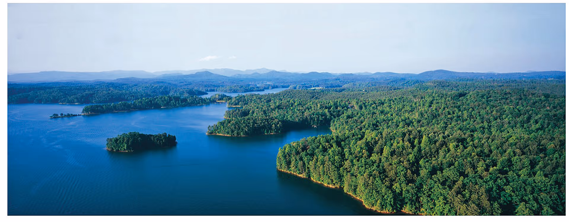 Aerial view of a large blue lake with forested shorelines and small tree-covered islands under a clear sky.