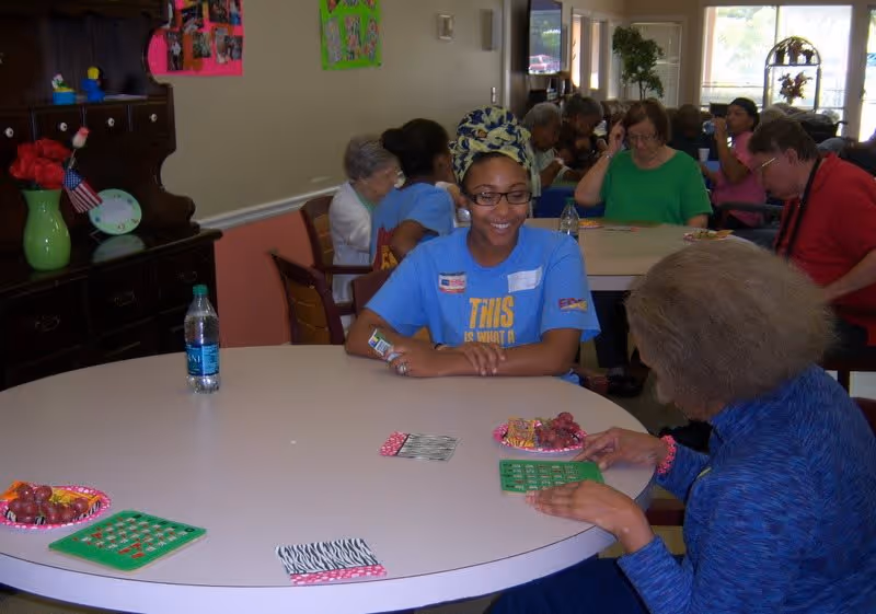 A group of elderly people and a caregiver sitting around tables playing bingo in a well-lit room with windows and decorations on the walls.